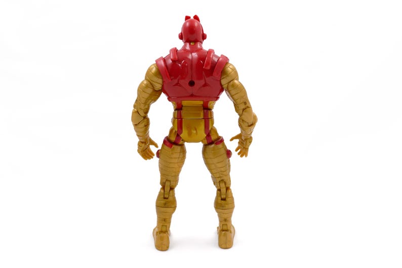 May include: Rear view of a red and gold action figure. The figure has a red helmet and chest piece, with gold-colored arms, legs, and torso. The figure is articulated and stands on a white surface.