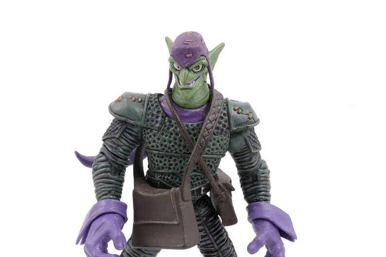 K&ouml;nnte beinhalten: Green Goblin Actionfigur mit lila Kappe, Handschuhen und Stiefeln. Die Figur hat einen finsteren Gesichtsausdruck und tr&auml;gt einen dunkelgr&uuml;nen Panzeranzug mit einer braunen Umh&auml;ngetasche. Sammlerst&uuml;ck.