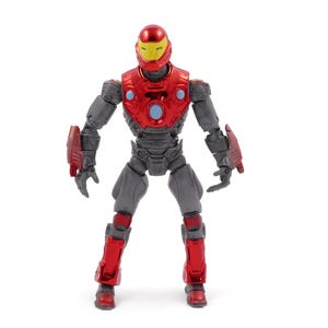 Op de afbeelding: Een rood en grijs actiefiguur van een superheld, tegen een witte achtergrond. Het figuur heeft een rode helm met een gele vizier, een rode borstplaat en rode accenten op de armen en benen. Het figuur is ongeveer 15 cm hoog.