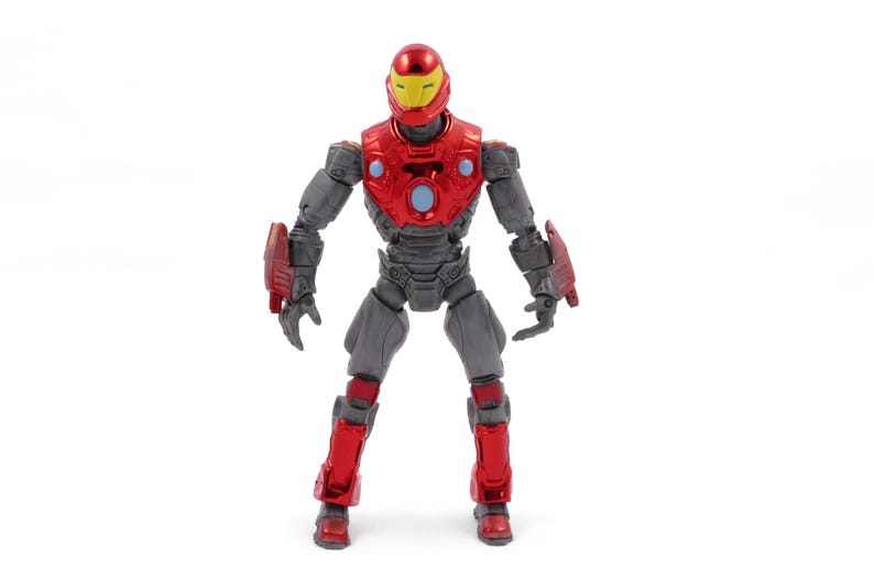 Op de afbeelding: Een rood en grijs actiefiguur van een superheld, tegen een witte achtergrond. Het figuur heeft een rode helm met een gele vizier, een rode borstplaat en rode accenten op de armen en benen. Het figuur is ongeveer 15 cm hoog.