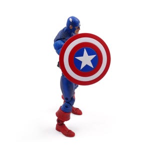 May include: A Captain America action figure in a blue and red suit, holding a shield with a white star on a blue circle, surrounded by red and white stripes. The figure is on a white background.