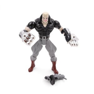 May include: A plastic action figure of a muscular character with a bald head, wearing a black top, gray pants, and brown boots. The figure has large silver gauntlets and a detached weapon on the ground.