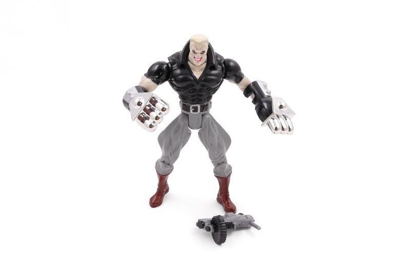 May include: A plastic action figure of a muscular character with a bald head, wearing a black top, gray pants, and brown boots. The figure has large silver gauntlets and a detached weapon on the ground.