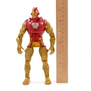 May include: A red and gold Iron Man action figure, approximately 7 inches tall, stands next to a wooden ruler. The figure has a red chest and helmet, with gold arms, legs, and torso. The figure is posed upright on a white background.