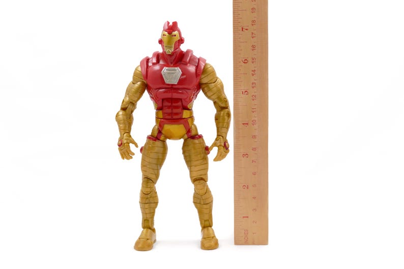May include: A red and gold Iron Man action figure, approximately 7 inches tall, stands next to a wooden ruler. The figure has a red chest and helmet, with gold arms, legs, and torso. The figure is posed upright on a white background.