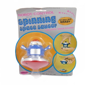 May include: A vintage toy robot space saucer with a blue and silver robot figure. The packaging says "Robot Control Spinning Space Saucer" and "Spins Like Crazy!"  The toy is made by Harmony and is wind-up.