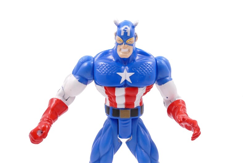 Pode incluir: Uma figura de a&ccedil;&atilde;o do Capit&atilde;o Am&eacute;rica em pose din&acirc;mica. A figura &eacute; predominantemente azul, com uma estrela branca no peito, listras vermelhas e brancas no torso e luvas vermelhas. A figura tem uma express&atilde;o severa e est&aacute; em um fundo branco.