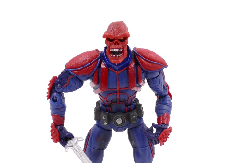 May include: A Red Skull action figure, a Marvel Comics supervillain, stands with a sword. The figure is predominantly blue and red, with a skull-like head and detailed armor. The figure is posed against a white background.