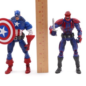 May include: Two action figures of comic book characters, Captain America and Red Skull, stand side-by-side. Captain America, in a blue, white, and red costume, holds a shield. Red Skull, in a red and blue outfit, holds a silver blade. A ruler is placed between them.