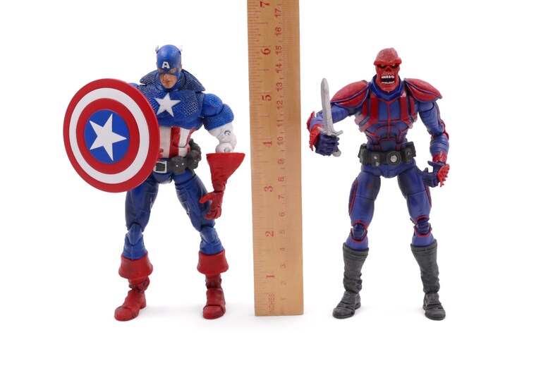 May include: Two action figures of comic book characters, Captain America and Red Skull, stand side-by-side. Captain America, in a blue, white, and red costume, holds a shield. Red Skull, in a red and blue outfit, holds a silver blade. A ruler is placed between them.