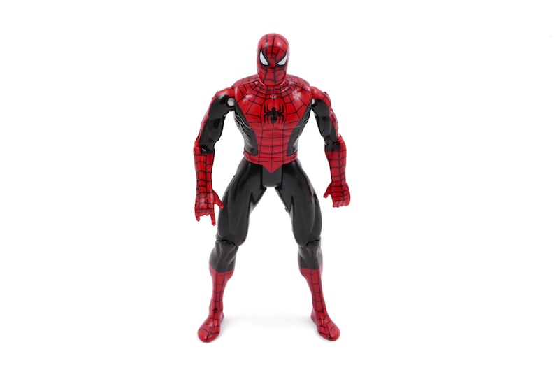 May include: A Spider-Man action figure in a standing pose. The figure is predominantly red and black, with a detailed web pattern on the red sections. The figure has a mask with white eye lenses.