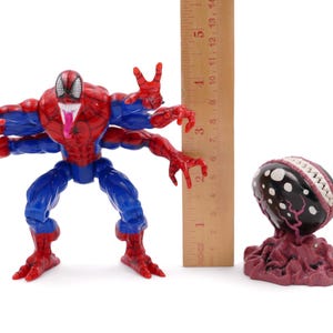 May include: A red, blue, and black Spider-Man action figure with four arms, a long tongue, and a wooden ruler. Next to it is a dark-colored, egg-shaped figure with white spots and a red base. The ruler measures up to 5 inches.