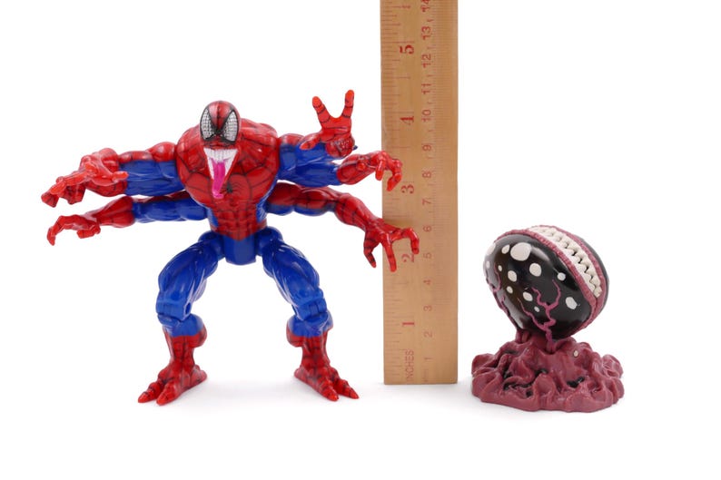 May include: A red, blue, and black Spider-Man action figure with four arms, a long tongue, and a wooden ruler. Next to it is a dark-colored, egg-shaped figure with white spots and a red base. The ruler measures up to 5 inches.