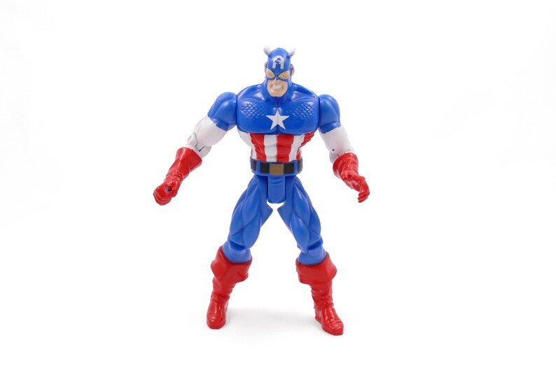 Pode incluir: Uma figura de a&ccedil;&atilde;o do Capit&atilde;o Am&eacute;rica em pose de p&eacute;. A figura &eacute; predominantemente azul, vermelha e branca, com uma estrela no peito. Possui um capacete azul com detalhes brancos e luvas e botas vermelhas. A figura tem cerca de 15 cm de altura.