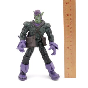 K&ouml;nnte beinhalten: Eine Green Goblin-Actionfigur, etwa 18 cm gro&szlig;, steht auf einer wei&szlig;en Oberfl&auml;che. Die Figur tr&auml;gt ein dunkelgr&uuml;nes und lila Kost&uuml;m mit einer Tasche und einem Hut. Ein Holzlineal befindet sich neben der Figur.