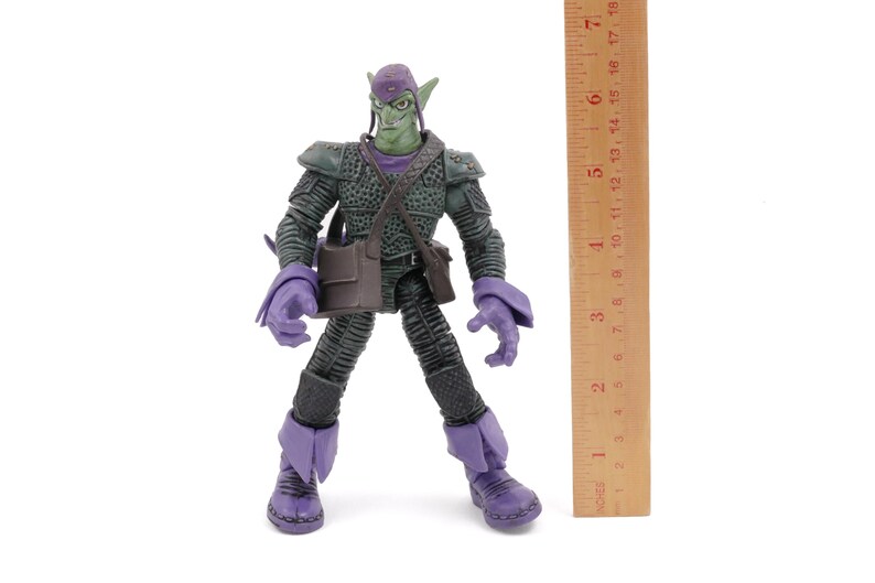 K&ouml;nnte beinhalten: Eine Green Goblin-Actionfigur, etwa 18 cm gro&szlig;, steht auf einer wei&szlig;en Oberfl&auml;che. Die Figur tr&auml;gt ein dunkelgr&uuml;nes und lila Kost&uuml;m mit einer Tasche und einem Hut. Ein Holzlineal befindet sich neben der Figur.