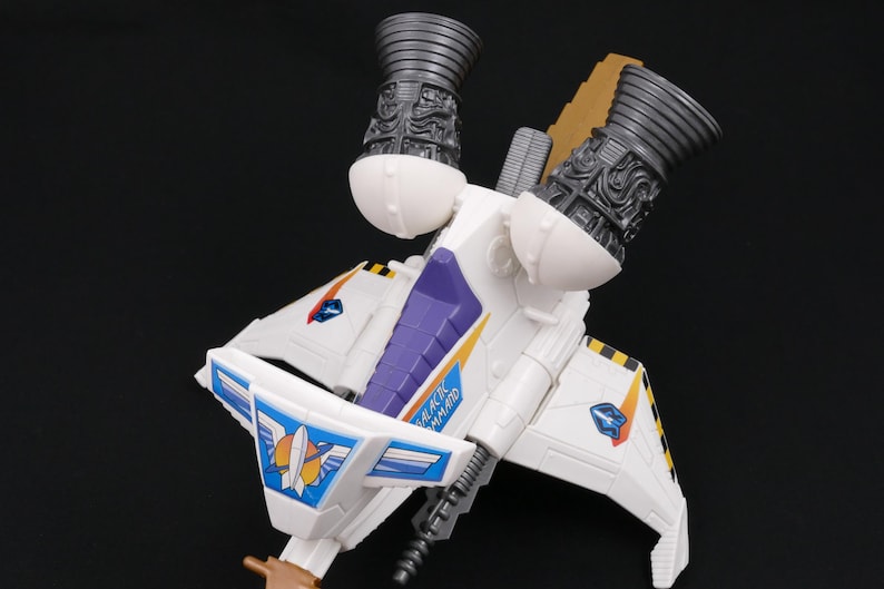 Op de afbeelding: Een wit speelgoed ruimteschip met paarse en gele accenten, met twee grote, gedetailleerde motoruitlaten. De vleugels hebben blauwe en oranje stickers. De tekst "GALACTIC COMMAND" is zichtbaar.