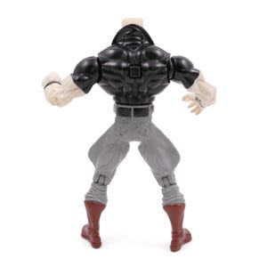 May include: A plastic action figure viewed from the back, featuring a black top, gray pants, and red boots. The figure has muscular arms and a neutral skin tone. The figure is posed with arms raised.
