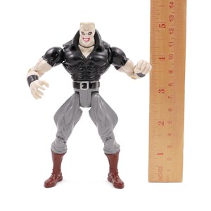 May include: A plastic action figure of a muscular character with a black top, gray pants, and brown boots. The figure has a pale complexion and a stern expression. A wooden ruler is positioned next to the figure, measuring approximately 5 inches.
