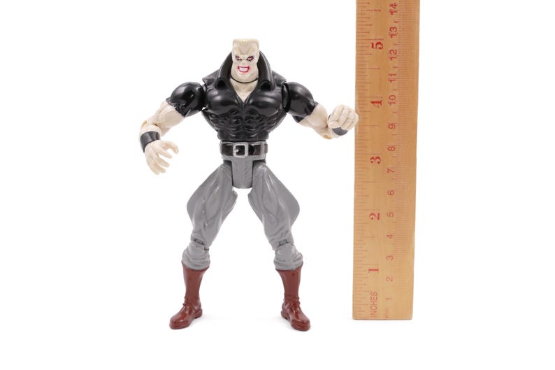 May include: A plastic action figure of a muscular character with a black top, gray pants, and brown boots. The figure has a pale complexion and a stern expression. A wooden ruler is positioned next to the figure, measuring approximately 5 inches.