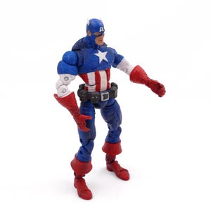 May include: A Captain America action figure in a dynamic pose. The figure is predominantly blue, red, and white, with a star emblem on the chest. It features a blue helmet, red gloves, and boots, and a black belt. The figure is approximately 6 inches tall.