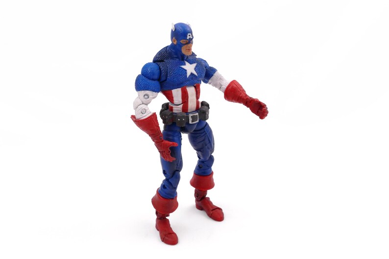 May include: A Captain America action figure in a dynamic pose. The figure is predominantly blue, red, and white, with a star emblem on the chest. It features a blue helmet, red gloves, and boots, and a black belt. The figure is approximately 6 inches tall.