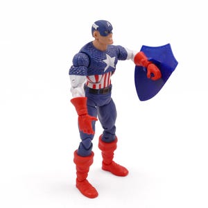 K&ouml;nnte beinhalten: Eine Captain America-Actionfigur. Die Figur tr&auml;gt ein blau-wei&szlig;es Kost&uuml;m mit einem Stern auf der Brust, einen blauen Helm und rote Handschuhe und Stiefel. Sie h&auml;lt einen blauen Schild.