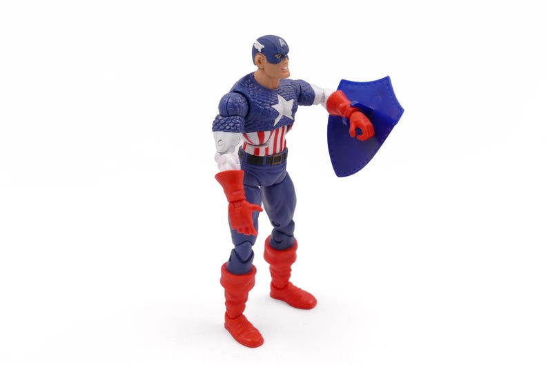 K&ouml;nnte beinhalten: Eine Captain America-Actionfigur. Die Figur tr&auml;gt ein blau-wei&szlig;es Kost&uuml;m mit einem Stern auf der Brust, einen blauen Helm und rote Handschuhe und Stiefel. Sie h&auml;lt einen blauen Schild.