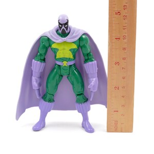 Puede incluir: Figura de acci&oacute;n verde con capa, botas y guantes morados. La figura tiene un emblema amarillo en el pecho y una m&aacute;scara. Una regla de madera mide aproximadamente 13 cm.