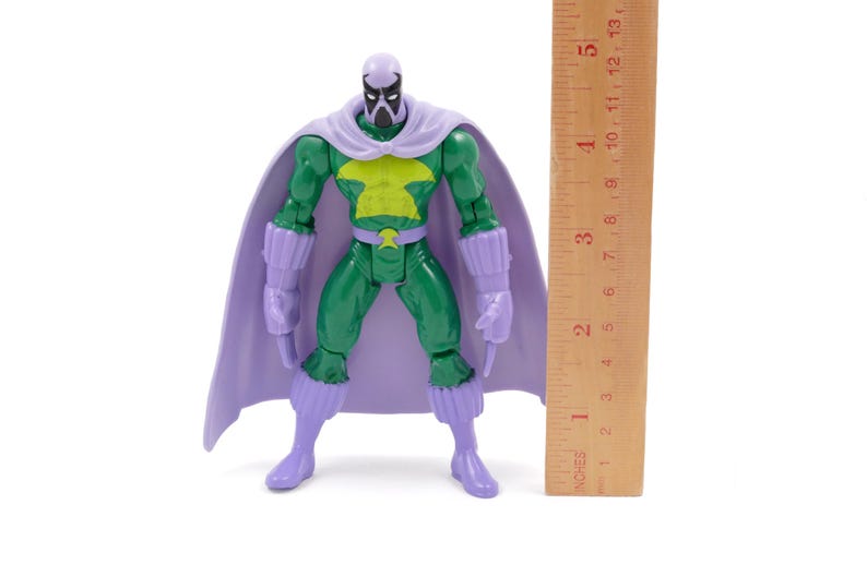 Puede incluir: Figura de acci&oacute;n verde con capa, botas y guantes morados. La figura tiene un emblema amarillo en el pecho y una m&aacute;scara. Una regla de madera mide aproximadamente 13 cm.