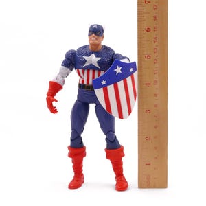 K&ouml;nnte beinhalten: Eine Captain America-Actionfigur, etwa 15 cm gro&szlig;, steht mit einem Schild und einem Lineal. Die Figur tr&auml;gt ein klassisches Kost&uuml;m mit blauem, wei&szlig;em und rotem Farbschema. Der Schild ist rot, wei&szlig; und blau.