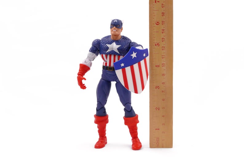 K&ouml;nnte beinhalten: Eine Captain America-Actionfigur, etwa 15 cm gro&szlig;, steht mit einem Schild und einem Lineal. Die Figur tr&auml;gt ein klassisches Kost&uuml;m mit blauem, wei&szlig;em und rotem Farbschema. Der Schild ist rot, wei&szlig; und blau.