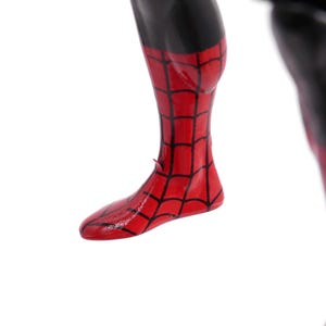 May include: Close-up of a red and black Spider-Man action figure's leg and foot. The red areas feature a black web pattern. The upper leg is black.