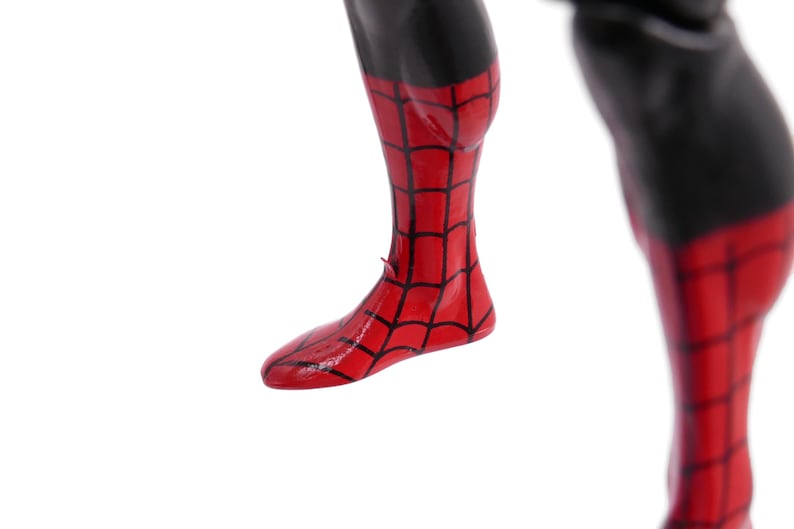 May include: Close-up of a red and black Spider-Man action figure's leg and foot. The red areas feature a black web pattern. The upper leg is black.