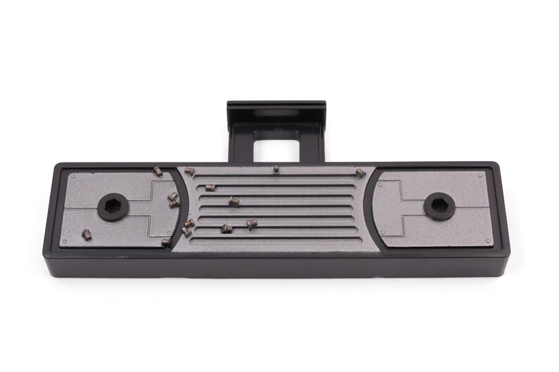 May include: A black and gray rectangular device with a raised section and a central slotted area. The device has two circular areas with a hexagonal center and several small, metallic objects scattered across the surface.