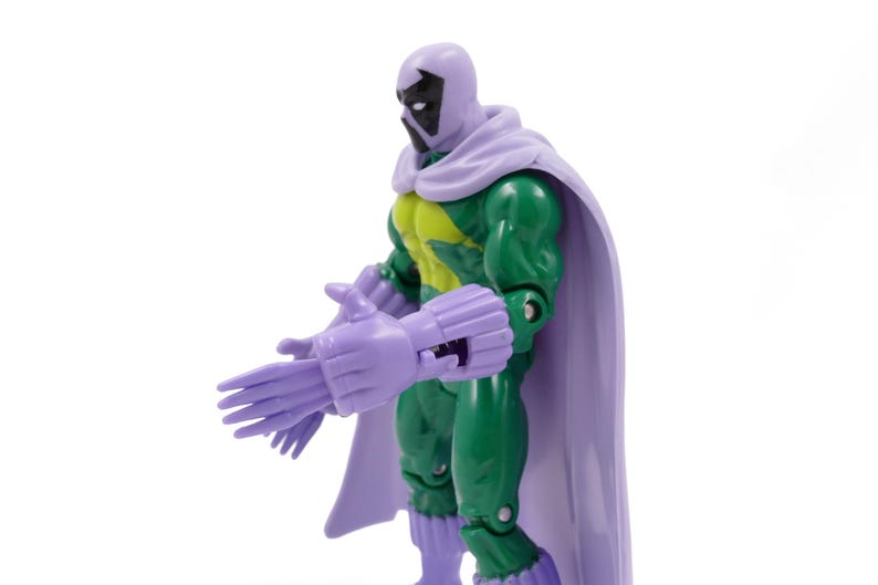 Puede incluir: Una figura de acci&oacute;n verde con capucha, capa y guantes morados. La figura tiene un torso amarillo y verde y detalles faciales negros. La figura posa con las manos extendidas.