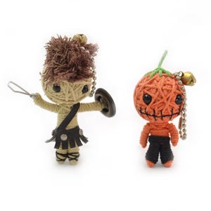 May include: Two handmade yarn dolls. One doll is a warrior with a shield and spear, wearing a black skirt and strap. The other is a pumpkin-shaped doll with a stitched face, wearing black pants. Both dolls have bells.