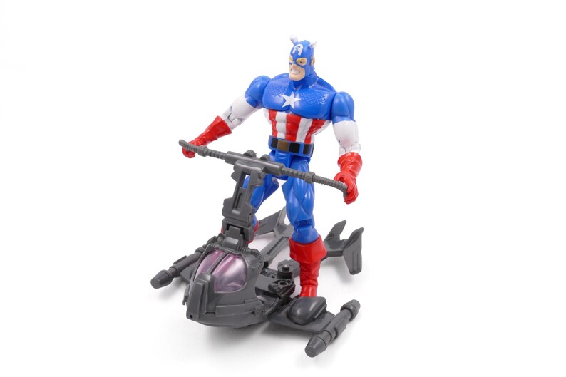 Pode incluir: Uma figura de a&ccedil;&atilde;o do Capit&atilde;o Am&eacute;rica em um traje azul, vermelho e branco, em p&eacute; em um ve&iacute;culo cinza com uma capota roxa. A figura tem uma estrela no peito e um capacete com asas.