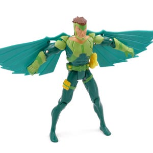 May include: A green and yellow action figure with teal wings. The figure is posed with its wings spread, showcasing a detailed design. It has a yellow pouch and a green belt. The figure is standing on a white surface.