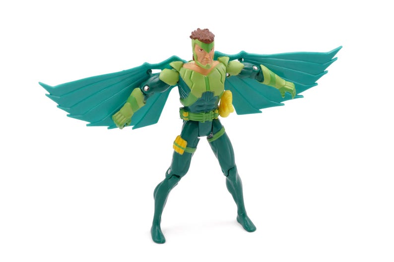 May include: A green and yellow action figure with teal wings. The figure is posed with its wings spread, showcasing a detailed design. It has a yellow pouch and a green belt. The figure is standing on a white surface.