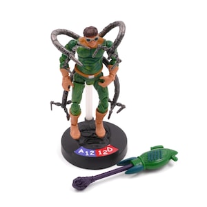 May include: A Dr. Octopus action figure in a green and brown outfit with four mechanical arms. The figure stands on a black base with red and blue text. A green and purple accessory lies nearby.
