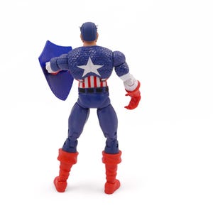 K&ouml;nnte beinhalten: Eine Captain America-Actionfigur, von hinten gesehen, steht auf einer wei&szlig;en Oberfl&auml;che. Die Figur tr&auml;gt ein blau-wei&szlig;es, sternenbesetztes Kost&uuml;m, rote Stiefel und Handschuhe und h&auml;lt einen blauen Schild. Auf dem R&uuml;cken der Figur ist ein gro&szlig;er wei&szlig;er Stern zu sehen.