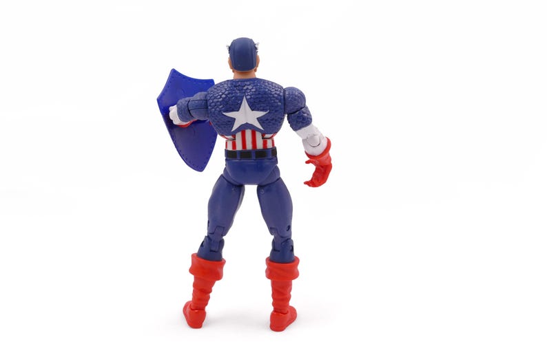 K&ouml;nnte beinhalten: Eine Captain America-Actionfigur, von hinten gesehen, steht auf einer wei&szlig;en Oberfl&auml;che. Die Figur tr&auml;gt ein blau-wei&szlig;es, sternenbesetztes Kost&uuml;m, rote Stiefel und Handschuhe und h&auml;lt einen blauen Schild. Auf dem R&uuml;cken der Figur ist ein gro&szlig;er wei&szlig;er Stern zu sehen.