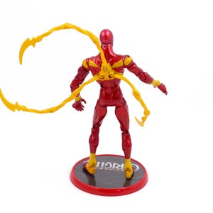 May include: Red and yellow action figure with four articulated arms. The figure is standing on a black base with the word "ORIGIN" printed on it.