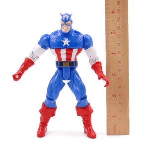 Pode incluir: Figura de a&ccedil;&atilde;o do Capit&atilde;o Am&eacute;rica em p&eacute;, com uma r&eacute;gua ao lado. A figura &eacute; principalmente azul, vermelha e branca, com uma estrela no peito. A r&eacute;gua mostra medidas em polegadas.