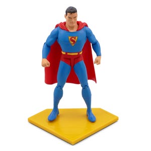 May include: A Superman action figure stands on a yellow hexagonal base. The figure wears a blue suit, red trunks, and a red cape. The Superman logo is on the chest. The figure has a determined expression.
