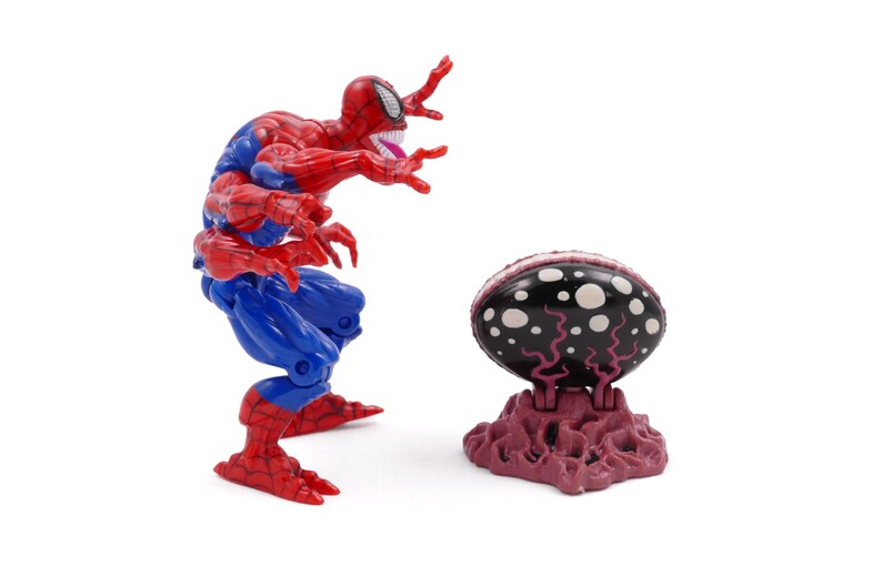 May include: A red and blue Spider-Man action figure with multiple arms and a menacing expression. Beside it is a black and white egg-shaped object on a purple base. The toy is likely a collectible.