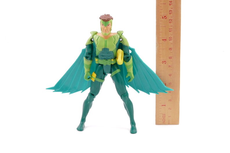 May include: A green and teal action figure of a superhero with a brown hairstyle and teal wings. The figure is wearing a green and teal suit with a yellow belt. A wooden ruler is next to the figure, measuring approximately 5 inches.