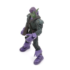 K&ouml;nnte beinhalten: Eine Green Goblin Actionfigur mit lila Helm, Handschuhen und Stiefeln. Die Figur tr&auml;gt einen dunkelgr&uuml;nen und grauen Anzug mit Schulterpolstern und einer braunen Umh&auml;ngetasche. Sie steht auf einer wei&szlig;en Oberfl&auml;che.