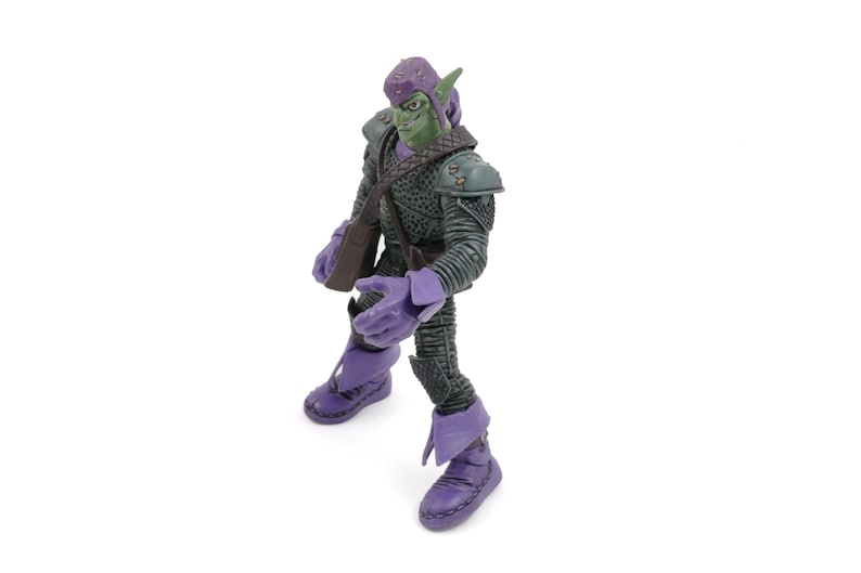 K&ouml;nnte beinhalten: Eine Green Goblin Actionfigur mit lila Helm, Handschuhen und Stiefeln. Die Figur tr&auml;gt einen dunkelgr&uuml;nen und grauen Anzug mit Schulterpolstern und einer braunen Umh&auml;ngetasche. Sie steht auf einer wei&szlig;en Oberfl&auml;che.